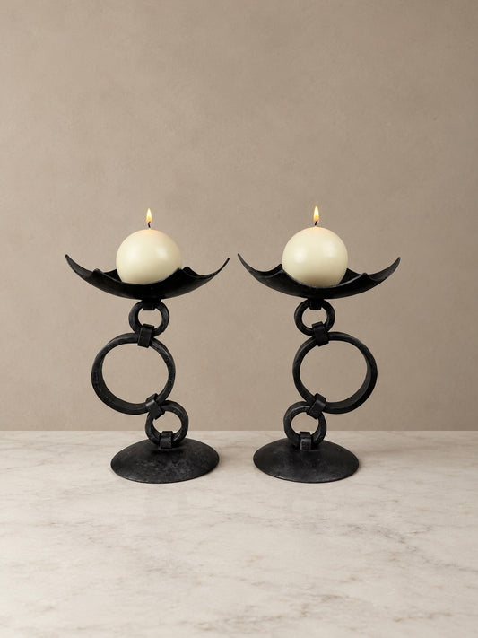 Pair of black wrought iron candlesticks with sculptural interlocking circular rings, curved candle dishes and round bases, each holding a lit ivory candle.